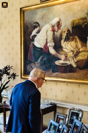 In London, a wedding photographer captures a candid moment at the reception venue. An older guest looks at framed photos on a table beneath a large portrait of an elderly man.